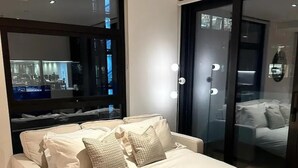 Ein modernes Schlafzimmer mit einem großen Bett, einer Glastür zu einem Badezimmer und einem großen Spiegel, der eine Stadtlandschaft reflektiert.