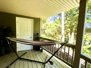 A hammock on a wooden deck with a white door and a view of greenery.
