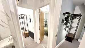 A hallway with a white door leading to a bedroom with a bed and a radiator. There is a small wooden stool and a black metal shelf with plants. - 6 Guests - 3 Bed House in Chester-le-street (Chester-le-Street)
