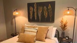 En pænt redt seng med et beige og brunstribet tæppe, to sengeborde med lamper og en væghængt billedkunst af fjer.