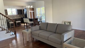 A spacious living room with a gray sofa, a glass coffee table, and a dining area with a kitchen in the background.