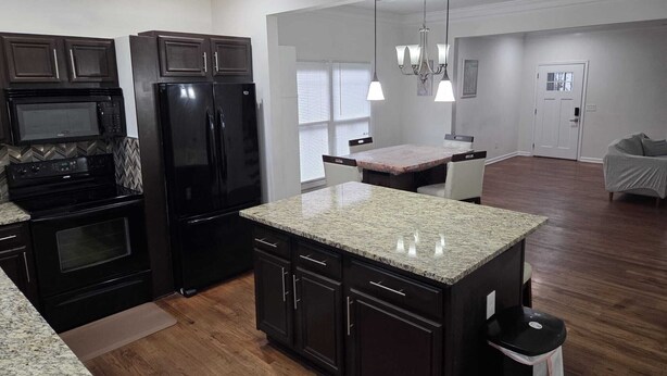 A modern kitchen with dark cabinetry, a granite countertop, a black refrigerator, and a microwave. There is a dining area with a table and chairs.