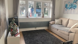 A living room with a beige sofa, a black rug, a small white side table with a Christmas tree, and a window with curtains. - Immaculate 2-bed Villa in Shrewsbury (Shrewsbury)