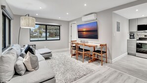 A modern living room with a sofa, a coffee table, a TV, and a dining area with chairs. - Lux Villa 3br 2ba Steps to Beach (Fort Lauderdale)