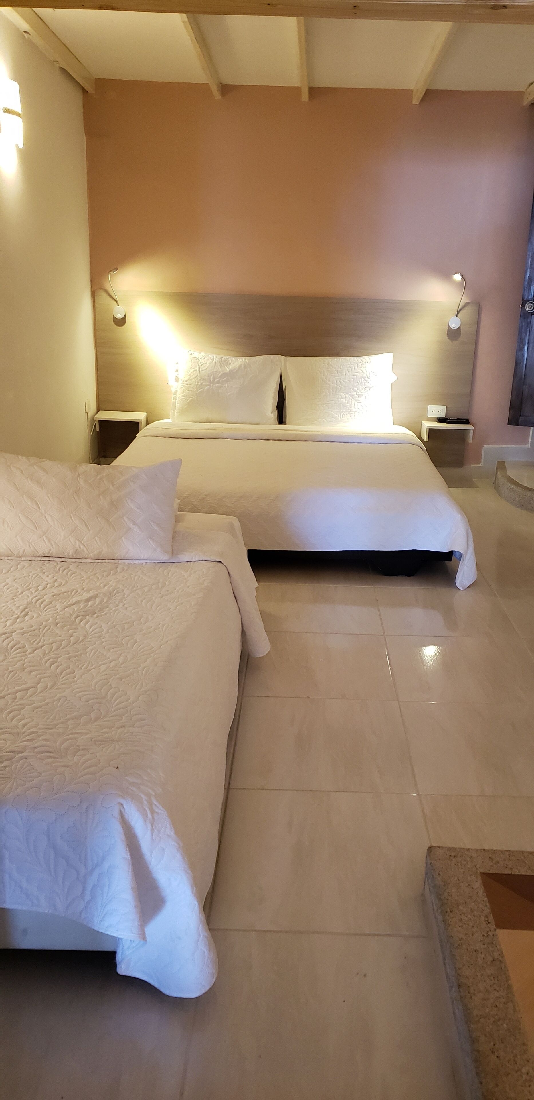 Habitación de hotel con dos camas, cabecero de madera, lámparas de noche y suelo de baldosas.