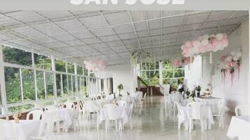 Un espace intérieur vaste et lumineux, avec des tables et des chaises blanches, de grandes fenêtres et un plafond orné d’un motif en réseau.