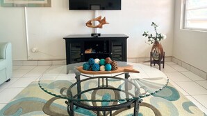 A living room with a flat-screen TV mounted on the wall, a glass coffee table with decorative items, a patterned rug, and a small plant. - New Listing! Island Bliss • 2BR • 3 Beds • 2 Baths (Saint Thomas)