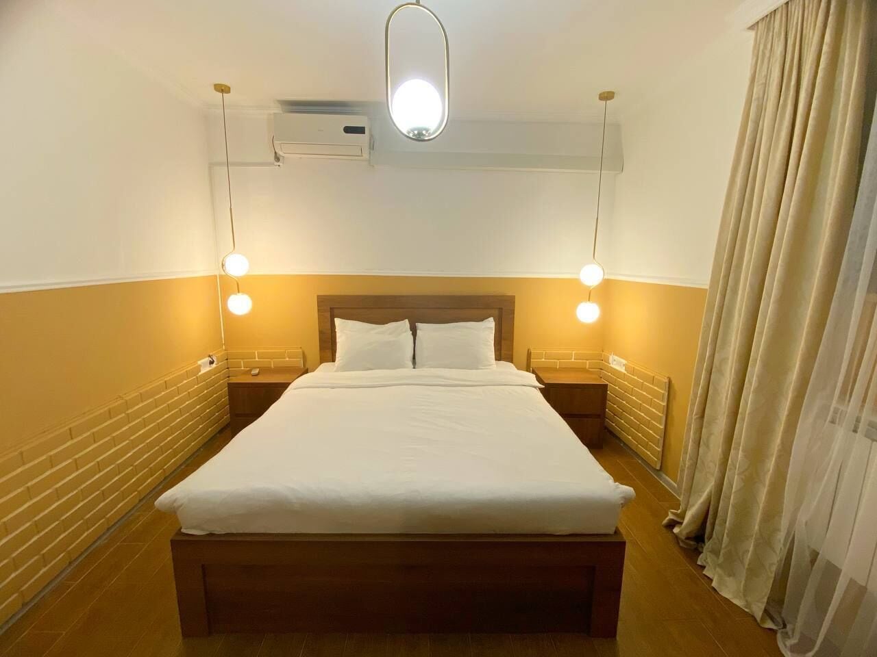 A hotel room with a wooden bed, two bedside tables, a wall-mounted air conditioner, and a ceiling light fixture.