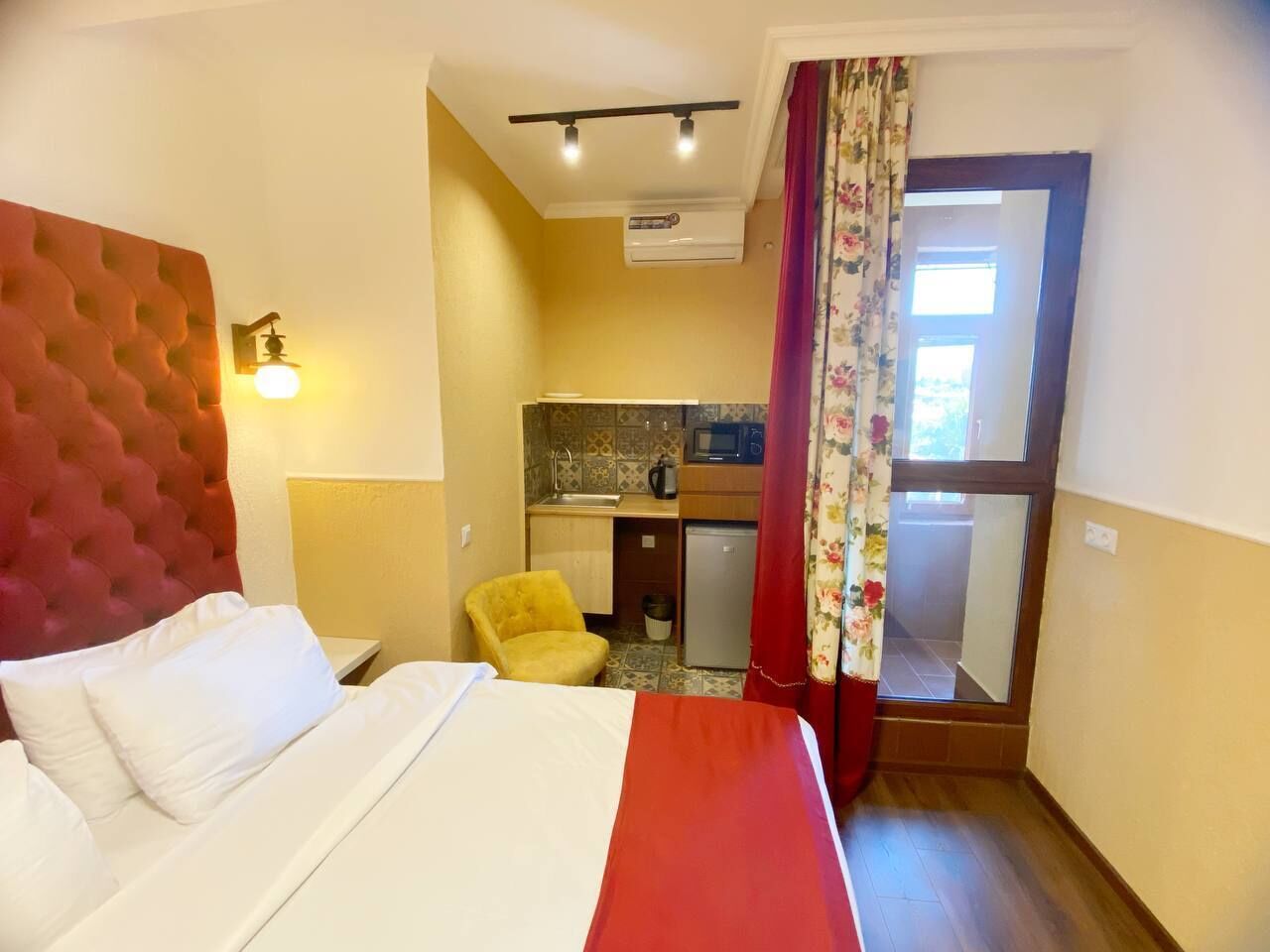 A hotel room with a bed, a small kitchenette, a mirror, and a window with curtains.