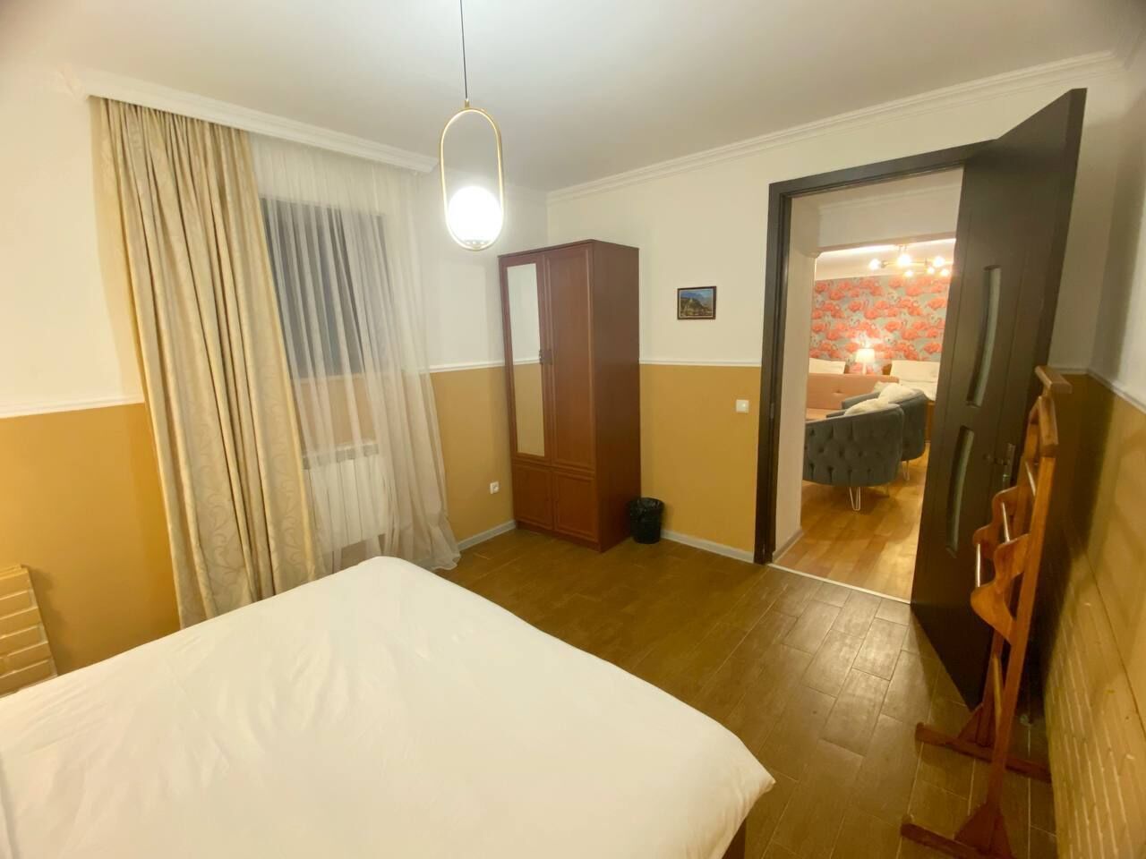 A hotel room with a bed, a wardrobe, a mirror, a door leading to a bathroom, and a small table with a lamp.