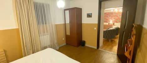 A hotel room with a bed, a wardrobe, a mirror, a door leading to a bathroom, and a small table with a lamp.