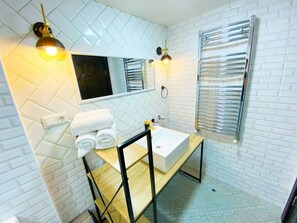 Een badkamer met een witte betegelde muur, een houten wastafelmeubel, een witte wasbak, een spiegel en twee wandlampen.