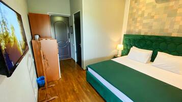 Habitación de hotel con cama verde, piso de madera, cabecera verde, aire acondicionado montado en la pared y una puerta que da a otra habitación.