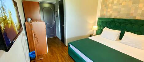 A hotel room with a green bed, wooden flooring, a green headboard, a wall-mounted air conditioner, and a door leading to another room.