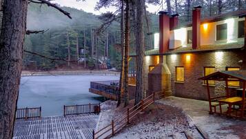 A lodge with a stone exterior, wooden deck, and a view of a frozen lake surrounded by trees.