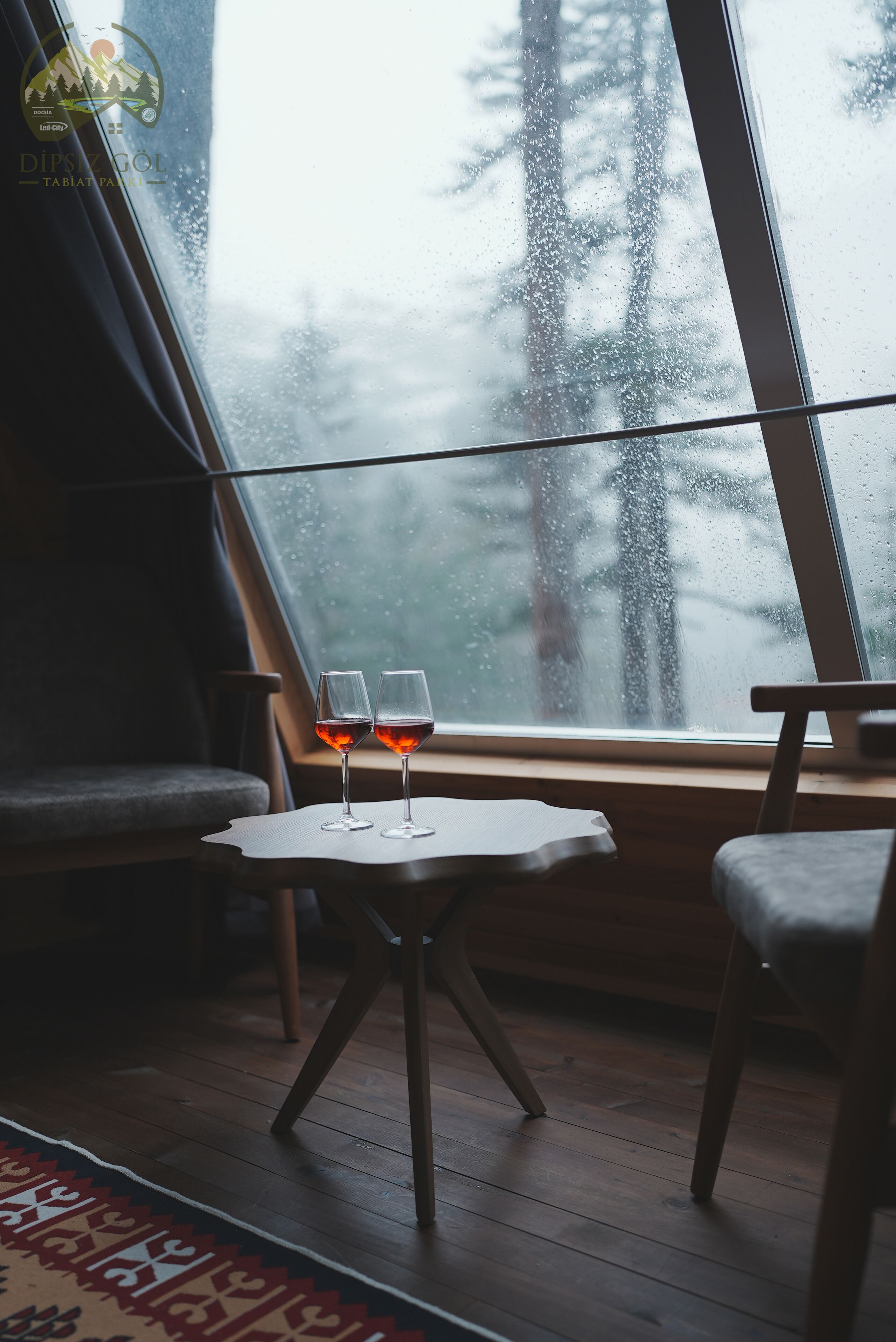 A cozy room with a large window, two glasses of red wine on a small table, and a chair.