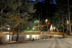 A snow-covered outdoor area with a wooden fence, a stone wall, and a building with illuminated windows. - Doceia Otel (Tosya)