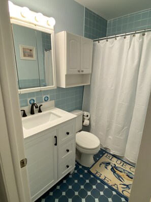 A bathroom with a white sink, mirror, and toilet, blue tiled walls, and a shower curtain. - Cape Roc Condo with Ocean Views and Heated Pool 1/2 Block to the Beach (Cape May)