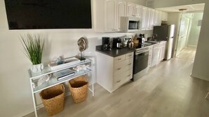 Eine moderne Küche mit weißen Schränken, einer Mikrowelle, einem Kühlschrank und einer Kaffeemaschine. Es gibt einen Glastisch mit Körben und einer dekorativen Fischskulptur.