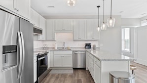 Una cocina moderna con armarios blancos, electrodomésticos de acero inoxidable, encimera de granito e iluminación colgante.