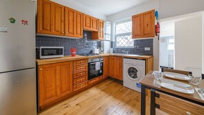 A compact kitchen with wooden cabinets, a microwave, a stove, a washing machine, and a dining table with a few items on it. - Modern Simplicity 10mins from Westfield, Stratford - Pass the Keys (London)