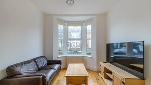 A living room with a brown leather sofa, a wooden coffee table, a TV on a wooden stand, and a bay window with a view of a house and street. - Modern Simplicity 10mins from Westfield, Stratford - Pass the Keys (London)