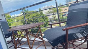A balcony with a wooden chair and a small table, offering a view of buildings and trees. - Apartment Sleeps 6 With Free Parking (Mombasa)