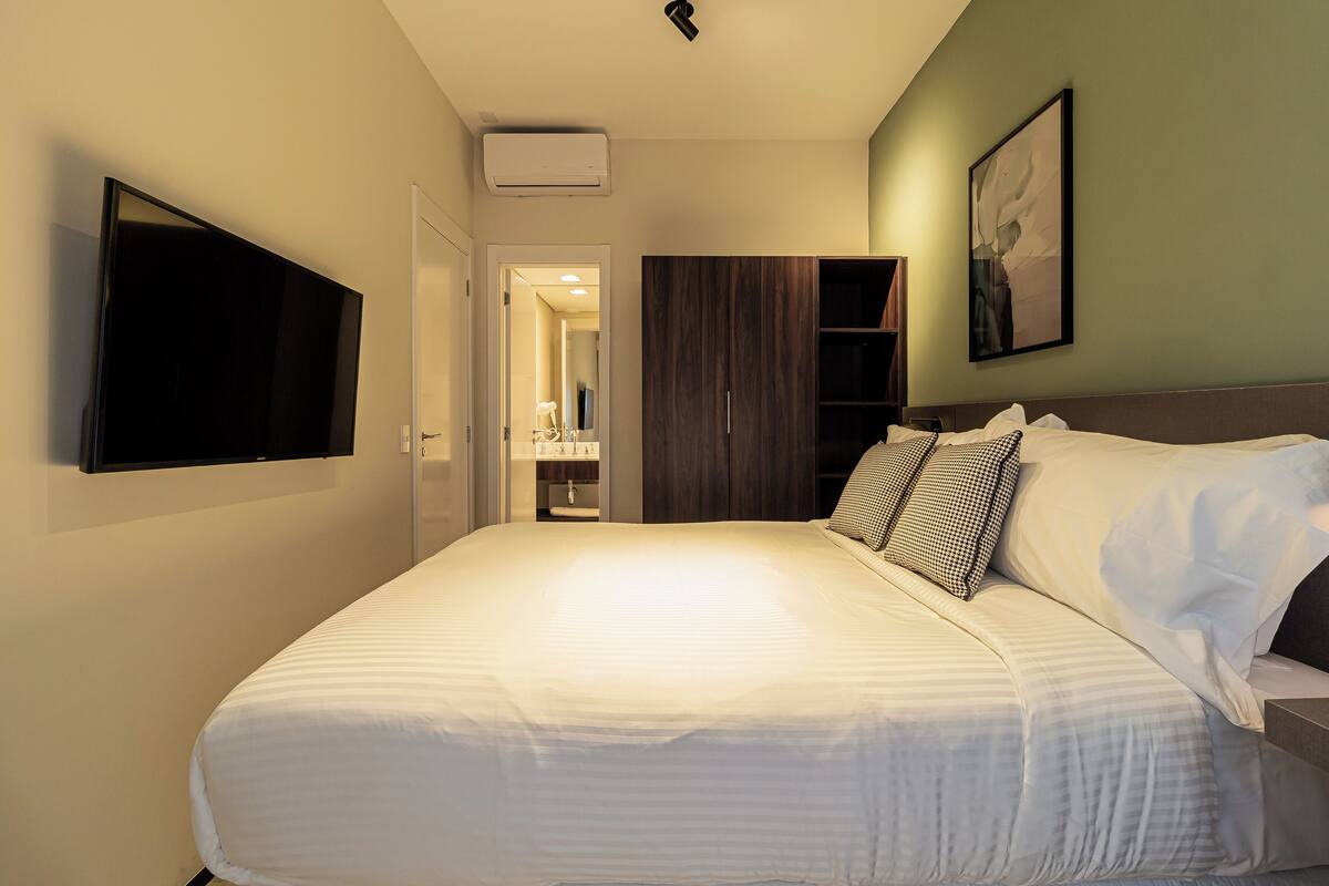 A hotel room with a large bed, a flat-screen TV mounted on the wall, a wardrobe, and a bathroom visible through an open door.