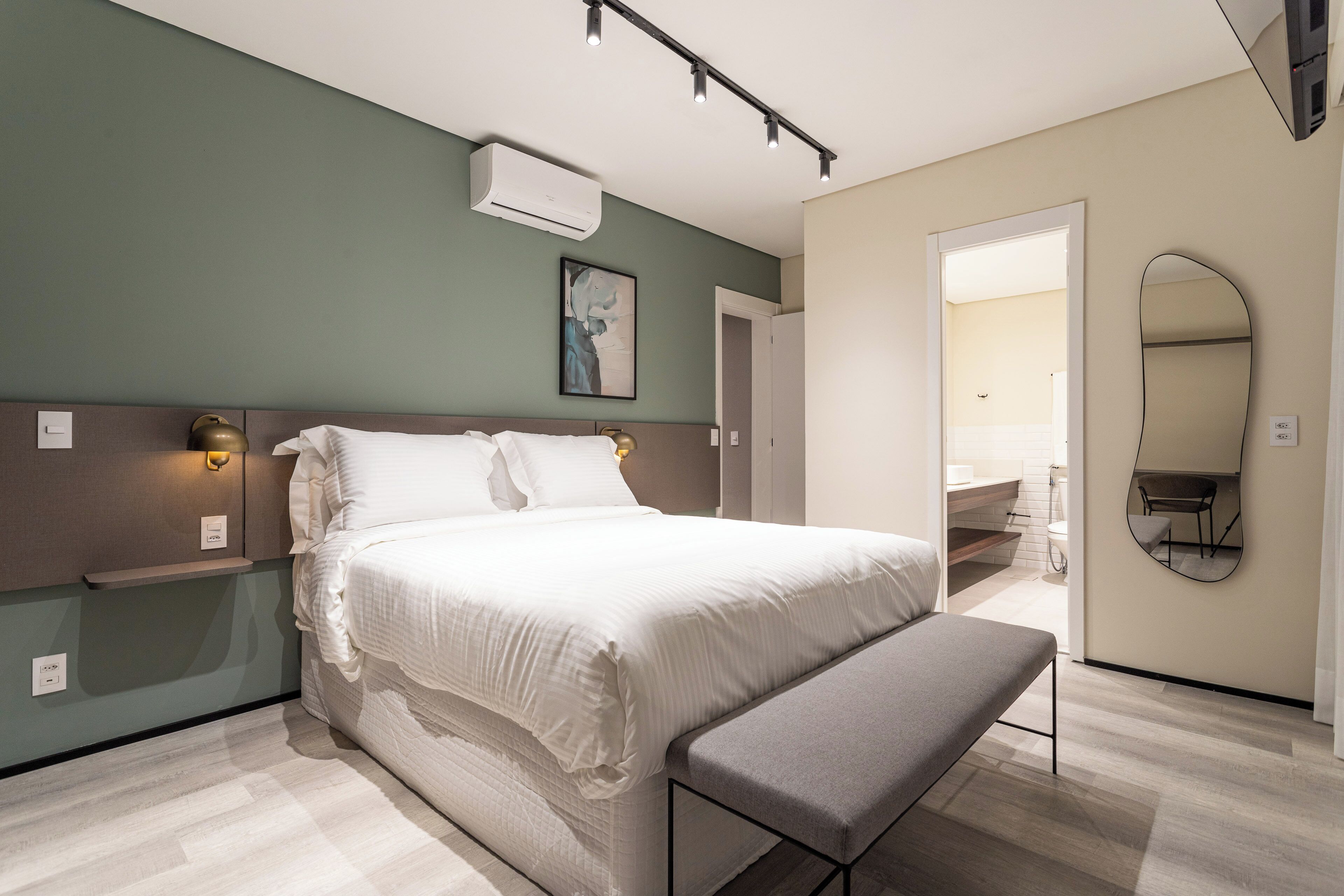 Une chambre moderne avec un grand lit, des tables de chevet, un banc, un miroir fixé au mur et une porte donnant sur une salle de bain.