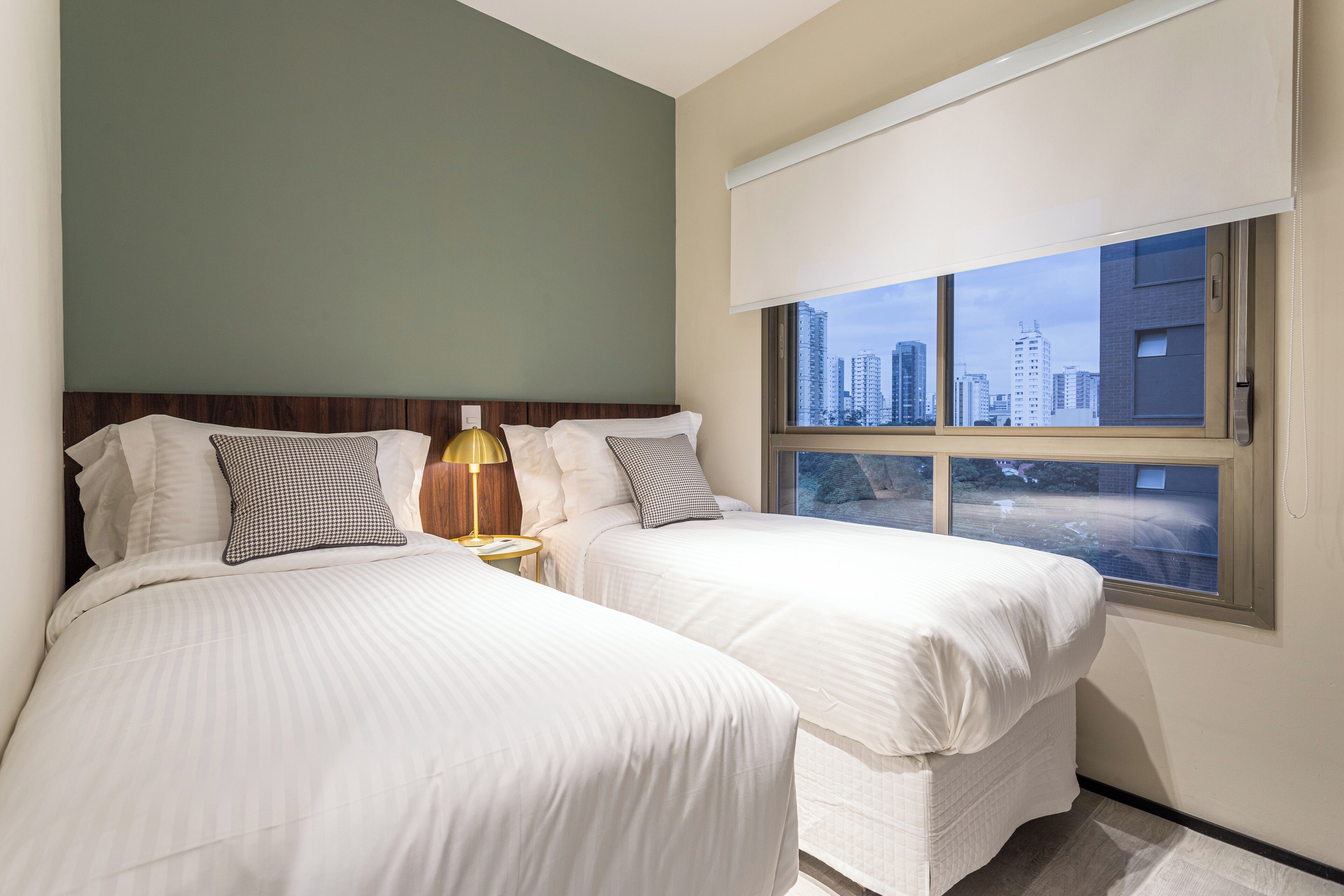A hotel room with two beds, a nightstand with a lamp, a window with a view of the cityscape, and a city skyline visible through the window.