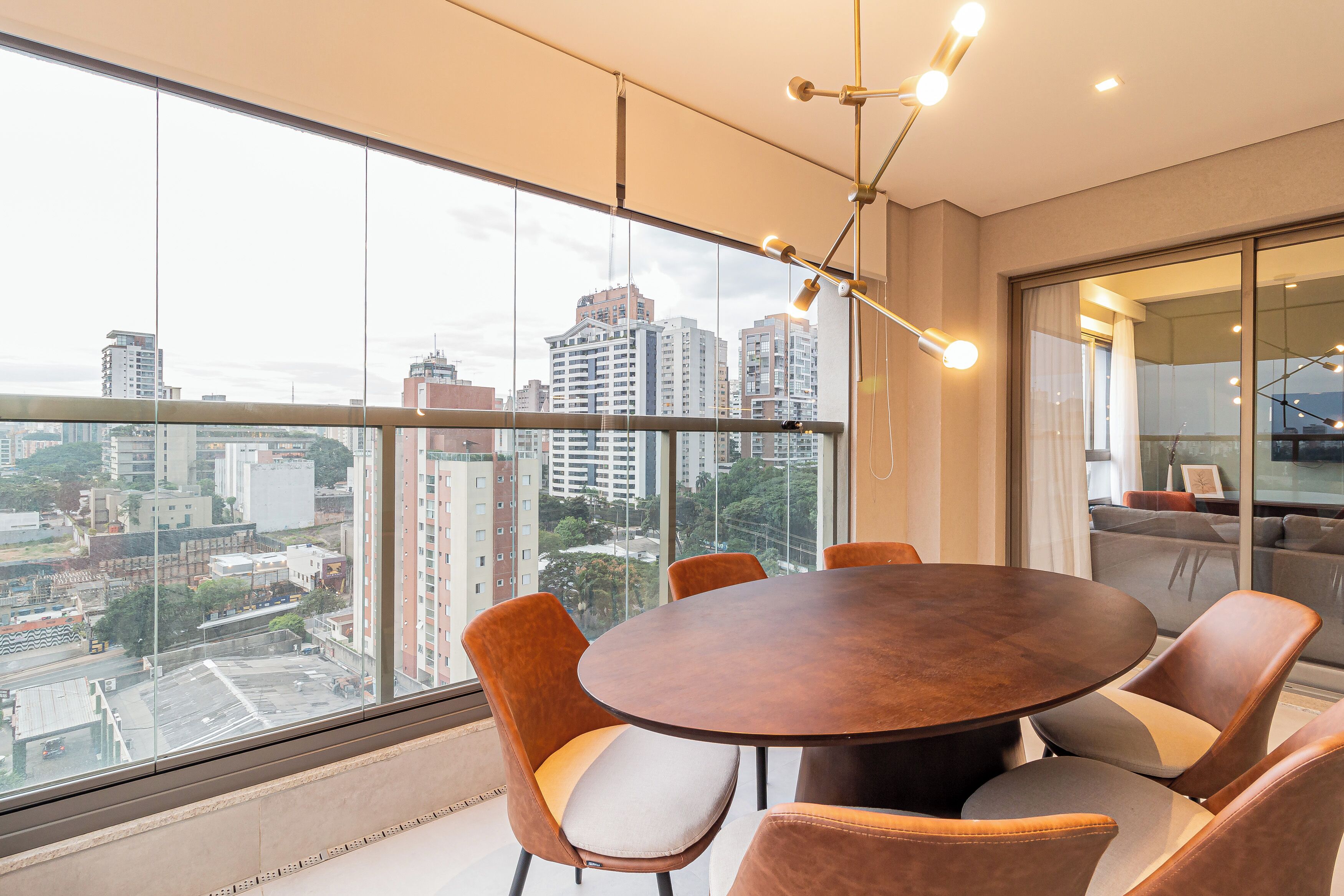 A modern dining area with a round table and cushioned chairs, large windows with a city view, and a contemporary chandelier.
