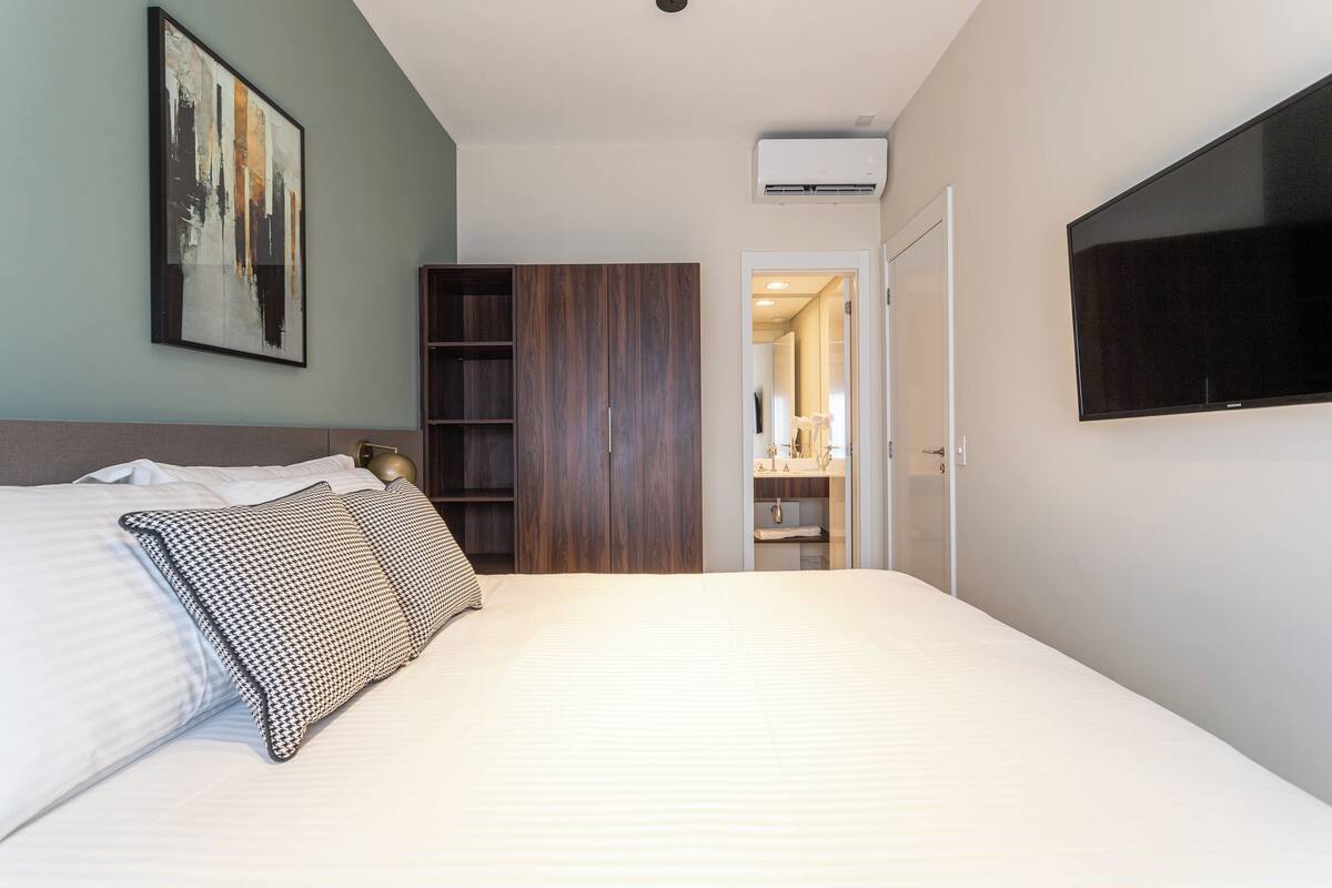 A modern hotel room with a large bed, a wardrobe, a TV, and an air conditioning unit.