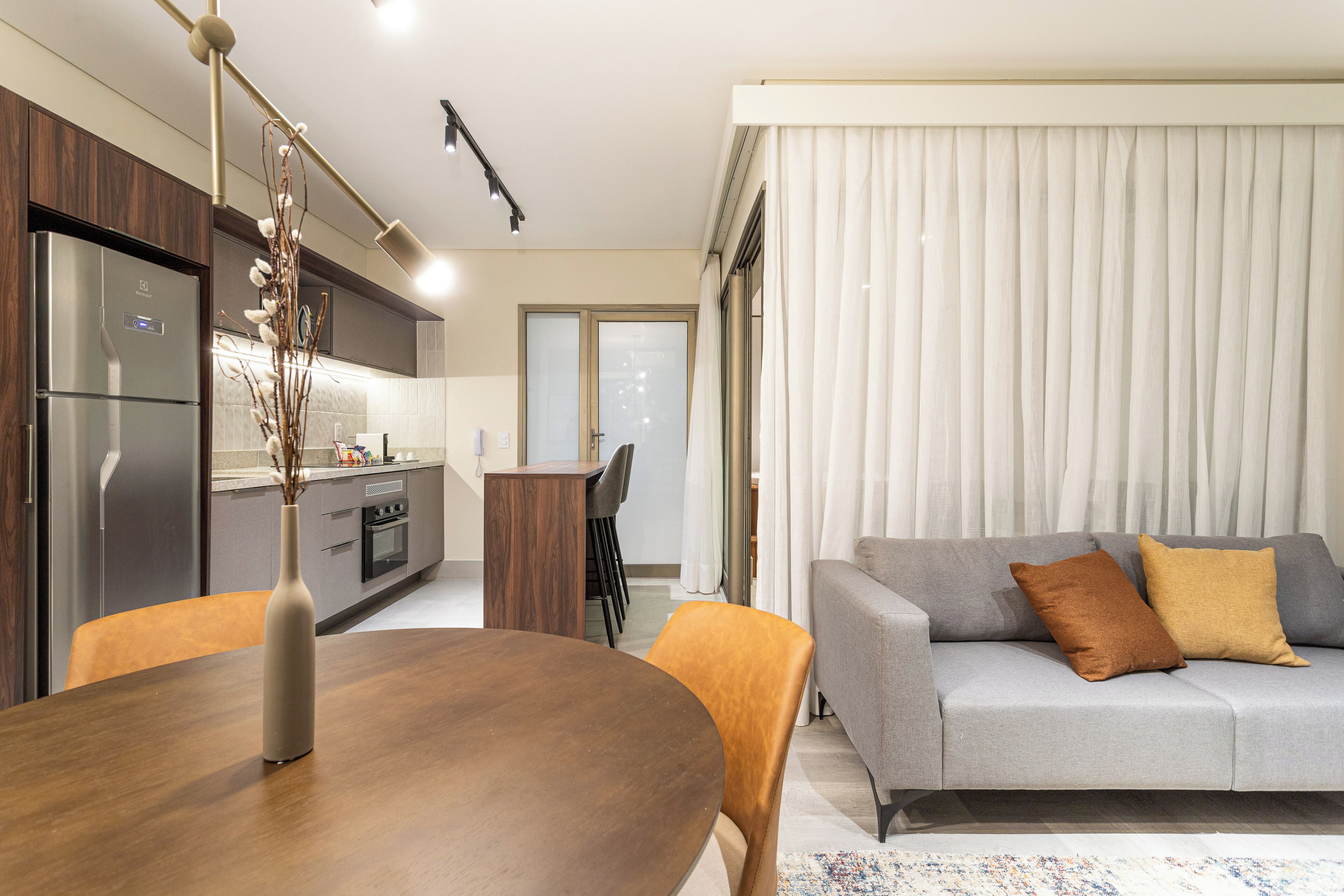 A modern kitchen with a built-in refrigerator, a dining table with chairs, and a grey sofa with cushions.