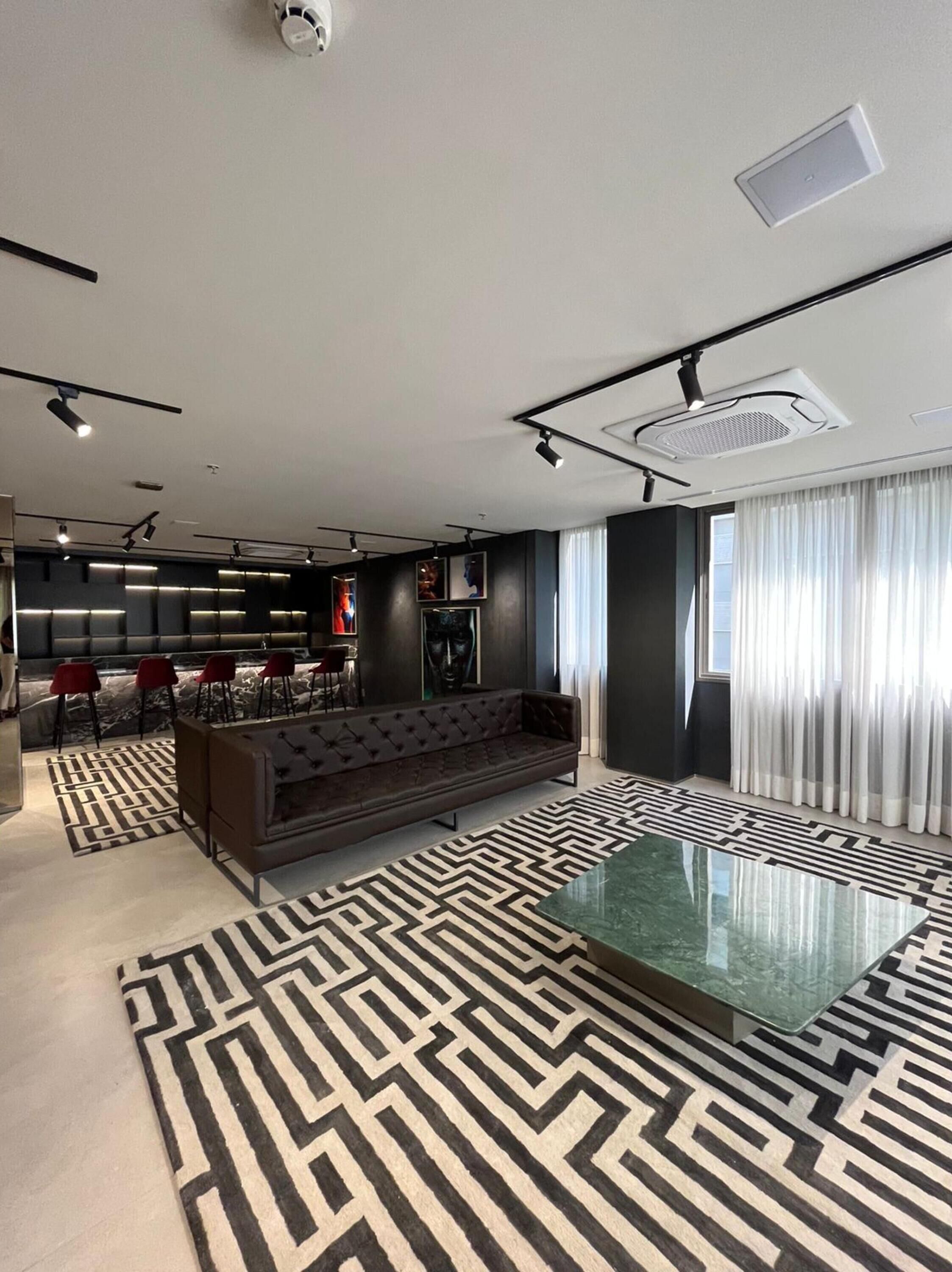 A modern living room with a sofa, a glass coffee table, and a patterned rug. There is a kitchen area in the background with red chairs and a marble countertop.