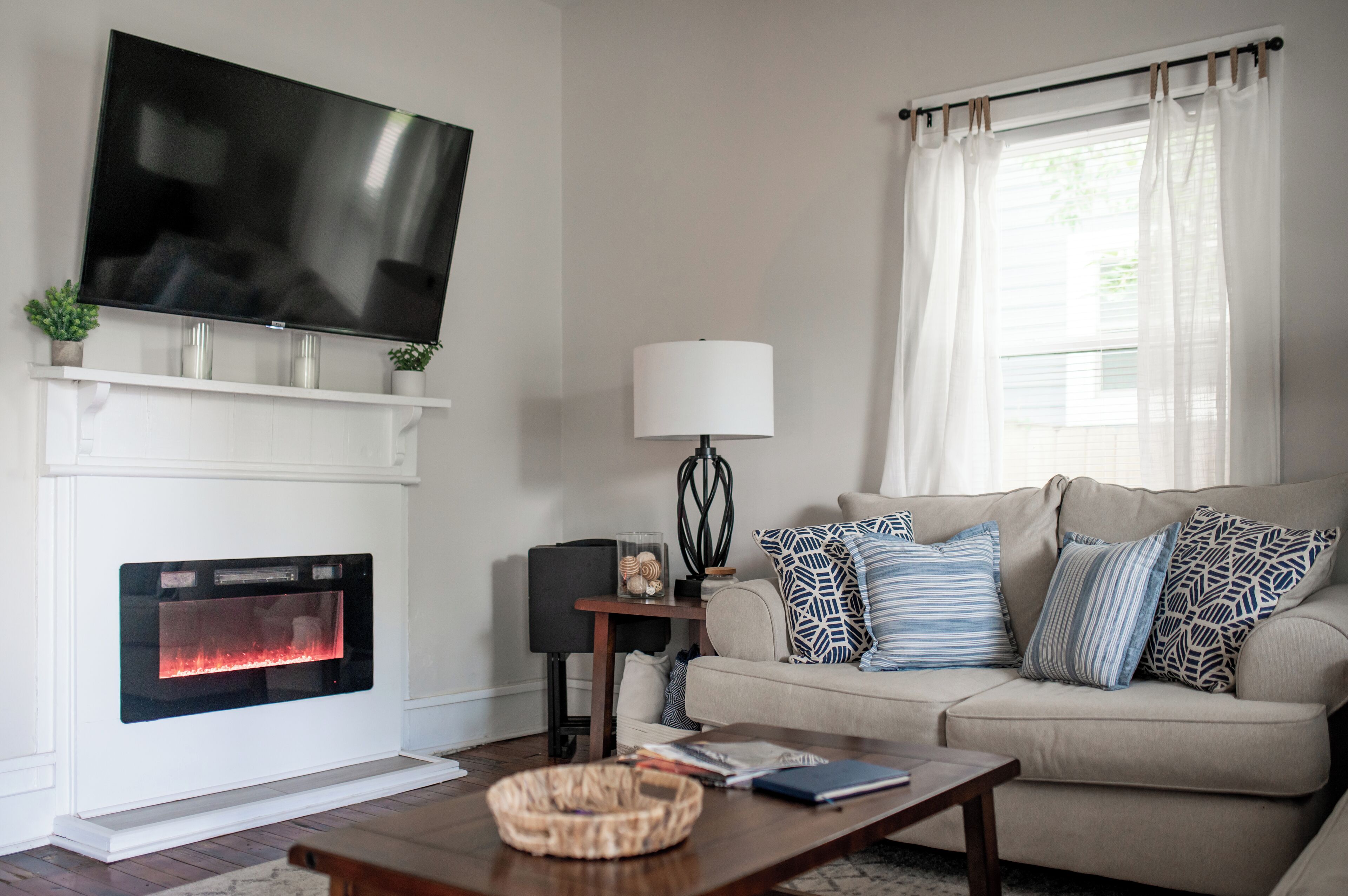 A living room with a fireplace, a television mounted above it, a sofa with patterned cushions, a lamp, and a coffee table.