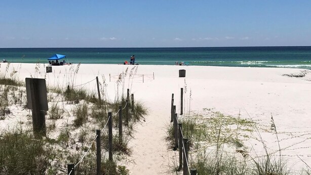 Une plage avec un ciel dégagé, un sentier sablonneux menant à l’eau, et quelques personnes et parasols.