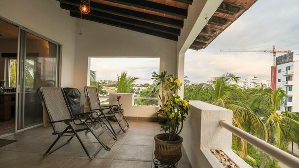 A balcony with two lounge chairs, a potted plant, and a view of a construction site. - Ocean View 2BR | Steps to Beach | Walk to Centro (Bucerías)