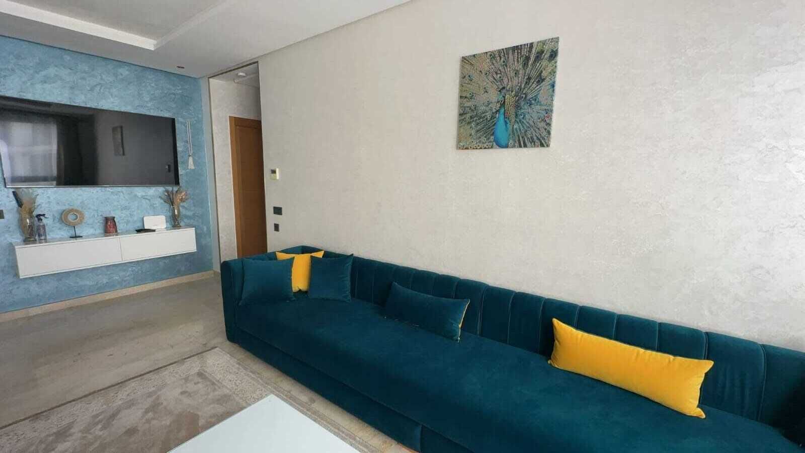 En moderne stue med en turkis sofa, gule puder, et vægmonteret fladskærms-tv og et dekorativt maleri på væggen.