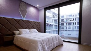 A bedroom with a large bed, a headboard, a nightstand, and a view of a building through a sliding door.