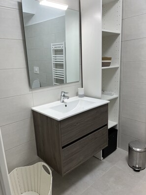 A modern bathroom with a white sink, wooden vanity, a large mirror, and a small trash bin. - Loc a Machja Fiurita 2 (Pietrosella)