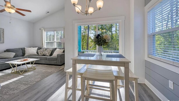 Un salon moderne avec un canapé gris, une table basse en bois et un coin repas comprenant une table et des chaises blanches. On y trouve également un ventilateur de plafond, un lustre et une fenêtre équipée de stores.