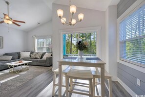 A modern living room with a grey sofa, a wooden coffee table, and a dining area with a white table and chairs. There is a ceiling fan, a chandelier, and a window with blinds. - Beautiful new home, 3 Bed 2.5 bath, 4min walk to beach (Myrtle Beach)