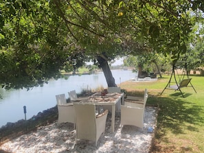 Un espace extérieur aménagé avec des chaises en osier et une table placés sous un arbre, près d’un lac.
