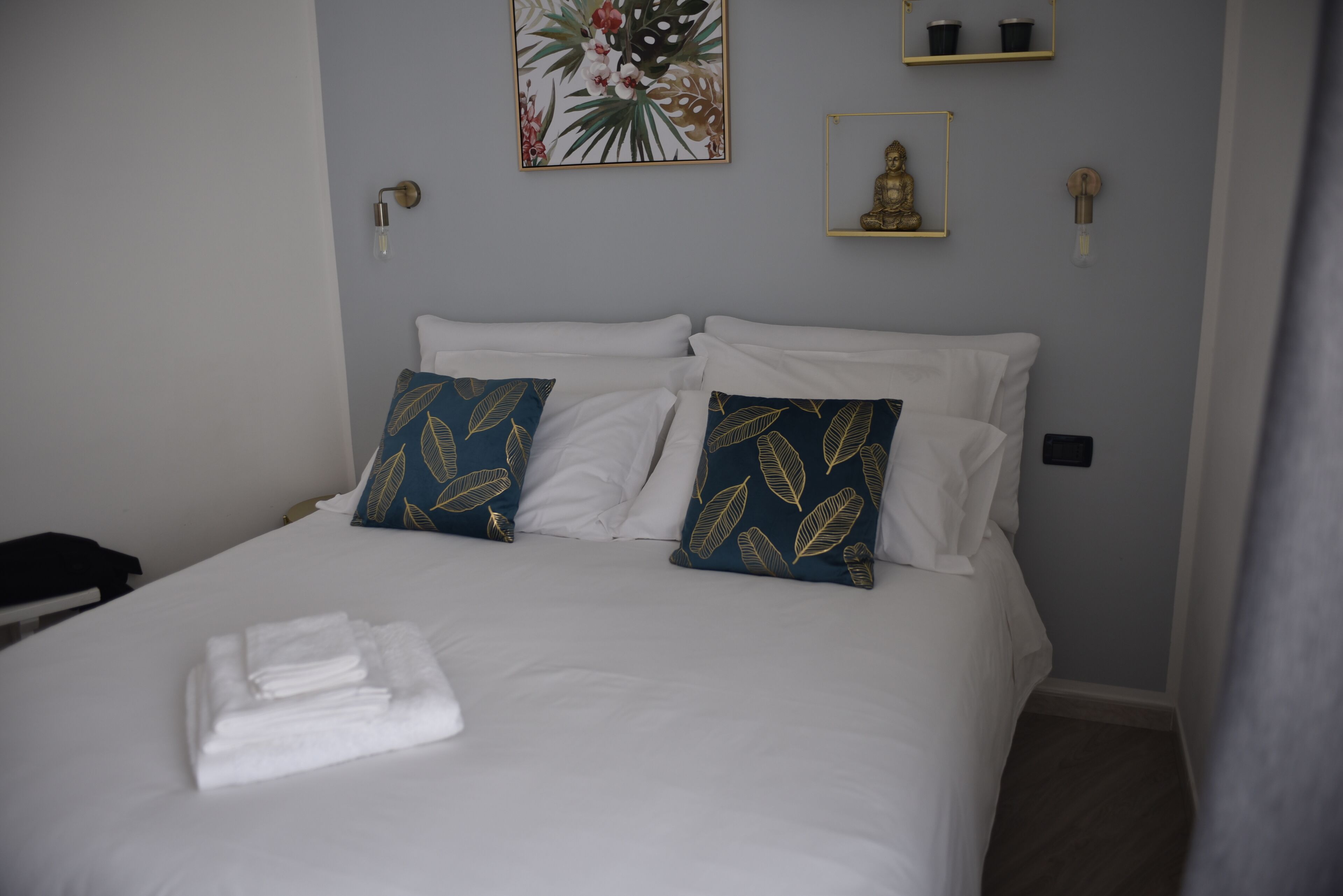 A neatly made bed with white linens and two decorative pillows, a wall-mounted light fixture, and a framed artwork above the bed.