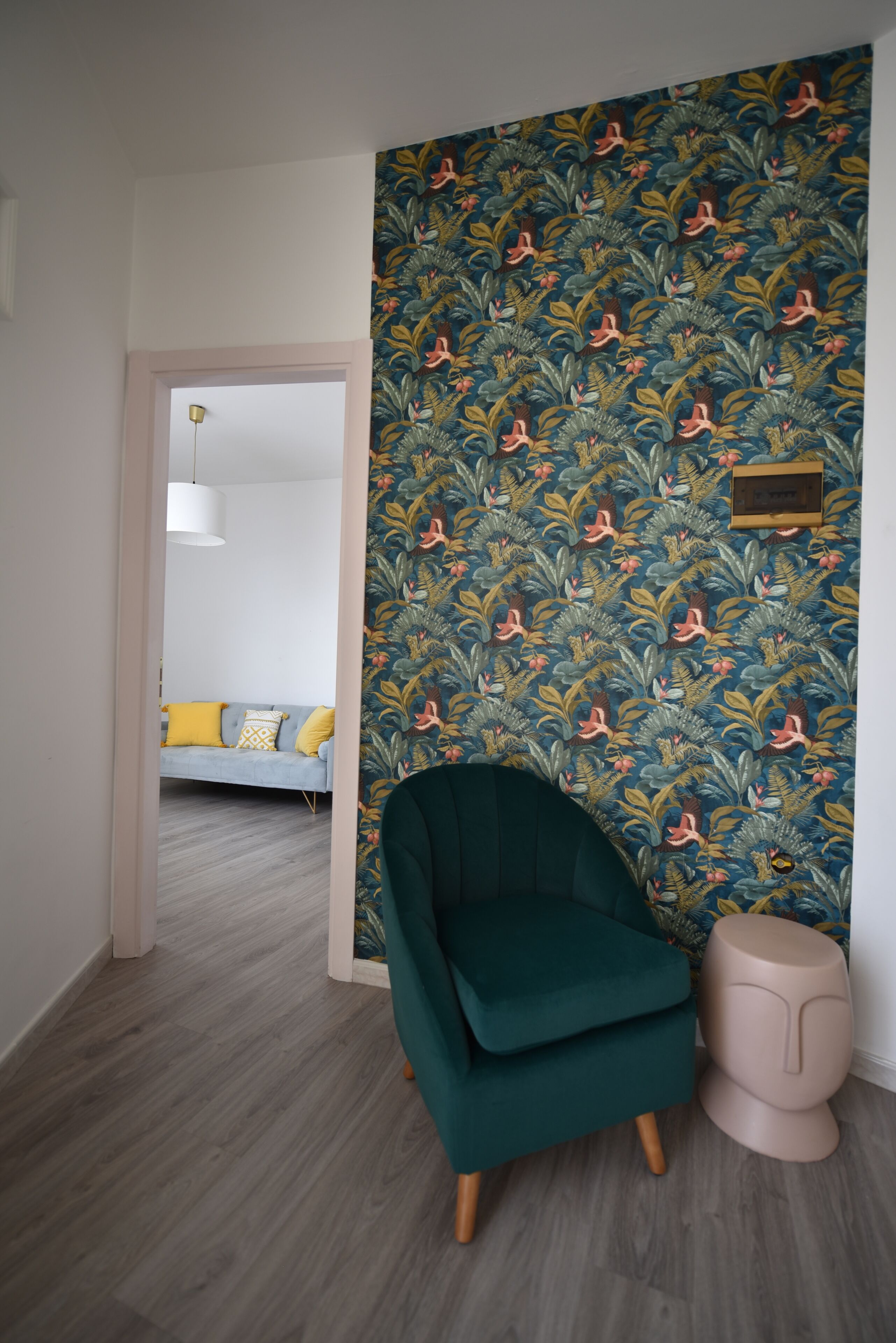 Una habitación con papel tapiz estampado, un sillón verde azulado, un pequeño taburete y un sofá con cojines amarillos al fondo.