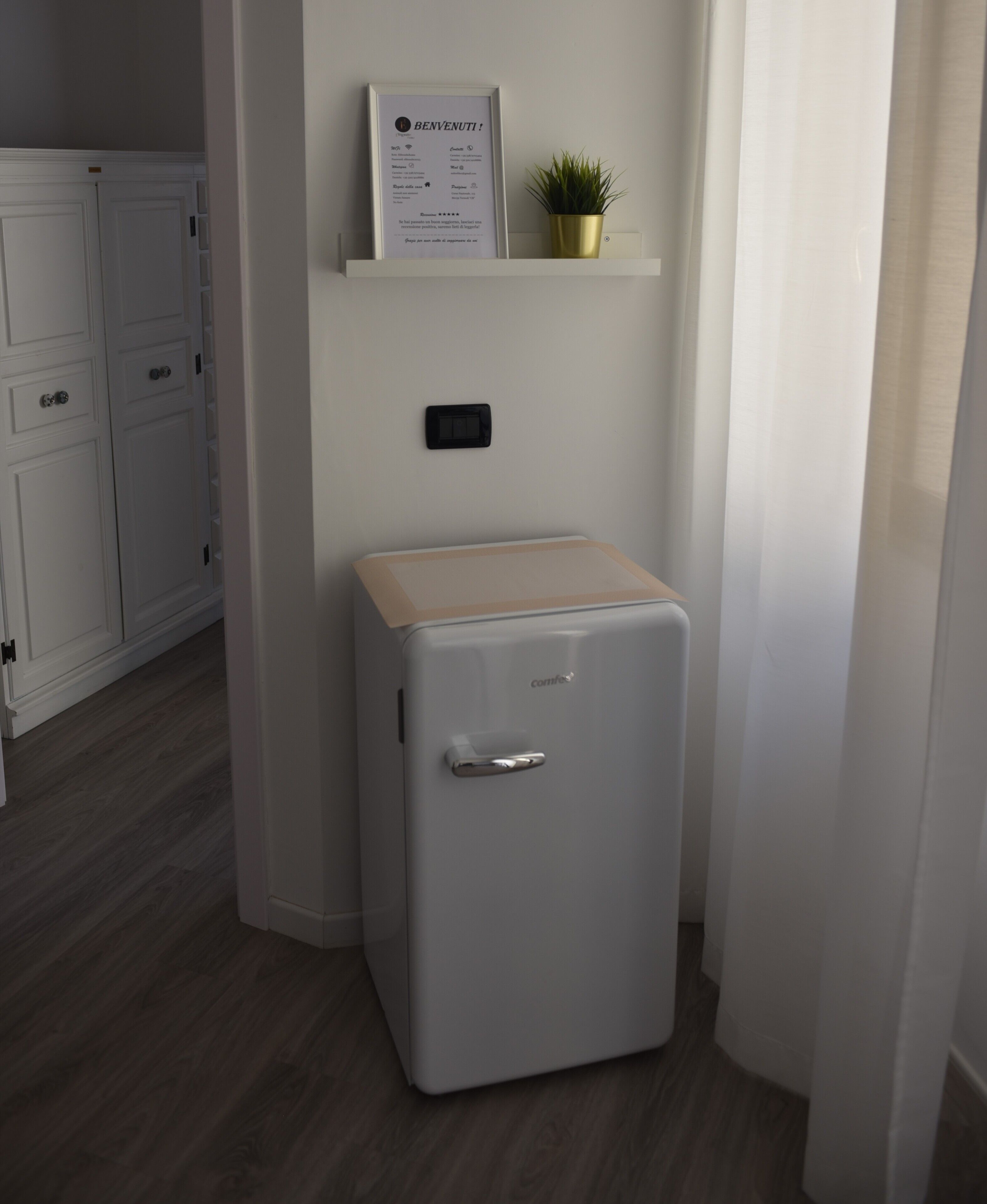 Un mini refrigerador compacto y blanco con una tapa de madera, una pequeña planta en maceta en un estante superior y un enchufe eléctrico negro en la pared.