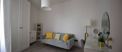 A modern bedroom with a grey sofa, white wardrobe, and a bedside table with a mirror and lamp.