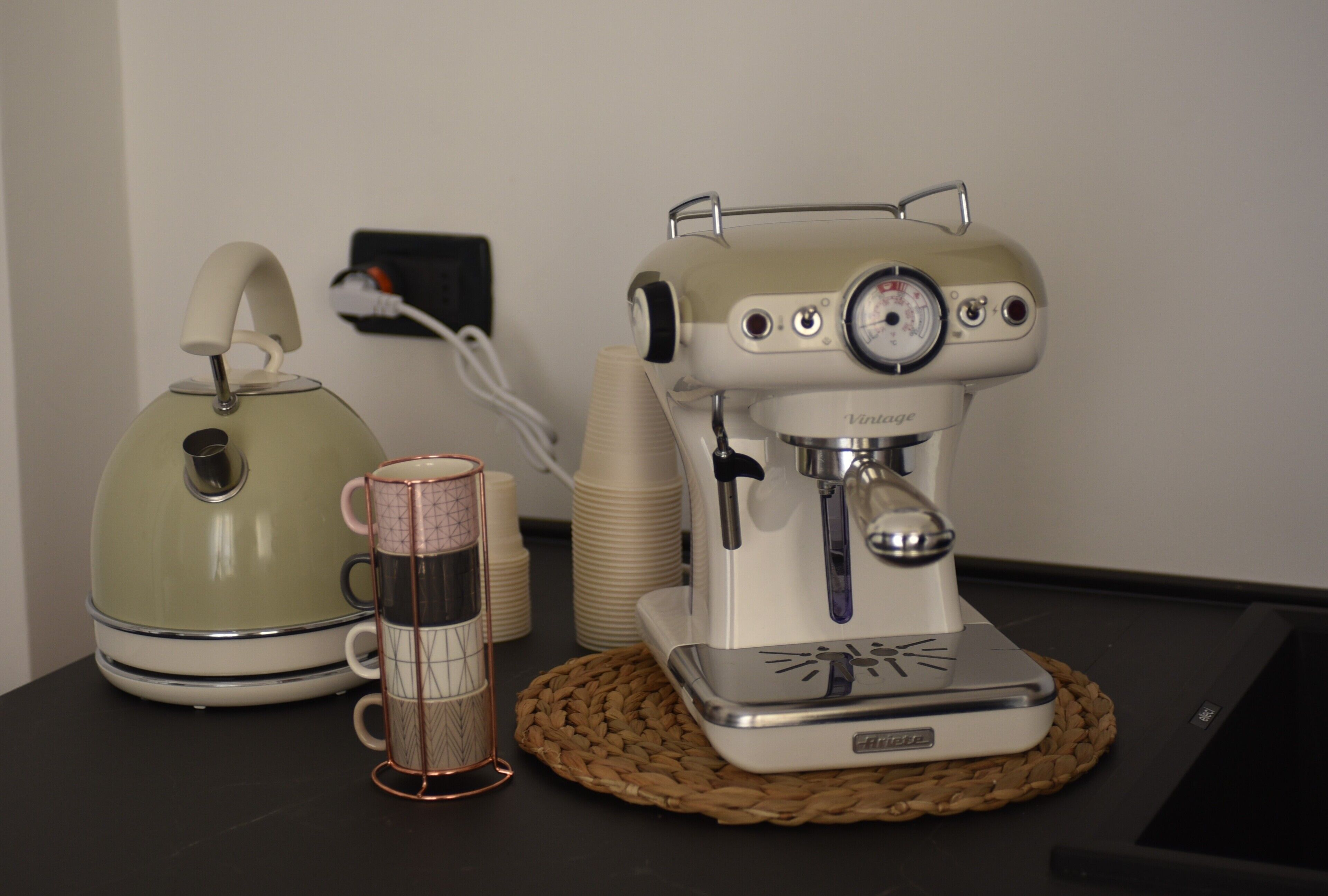 A vintage-style espresso machine on a woven mat, a green kettle, and a wire-framed mug on a table.
