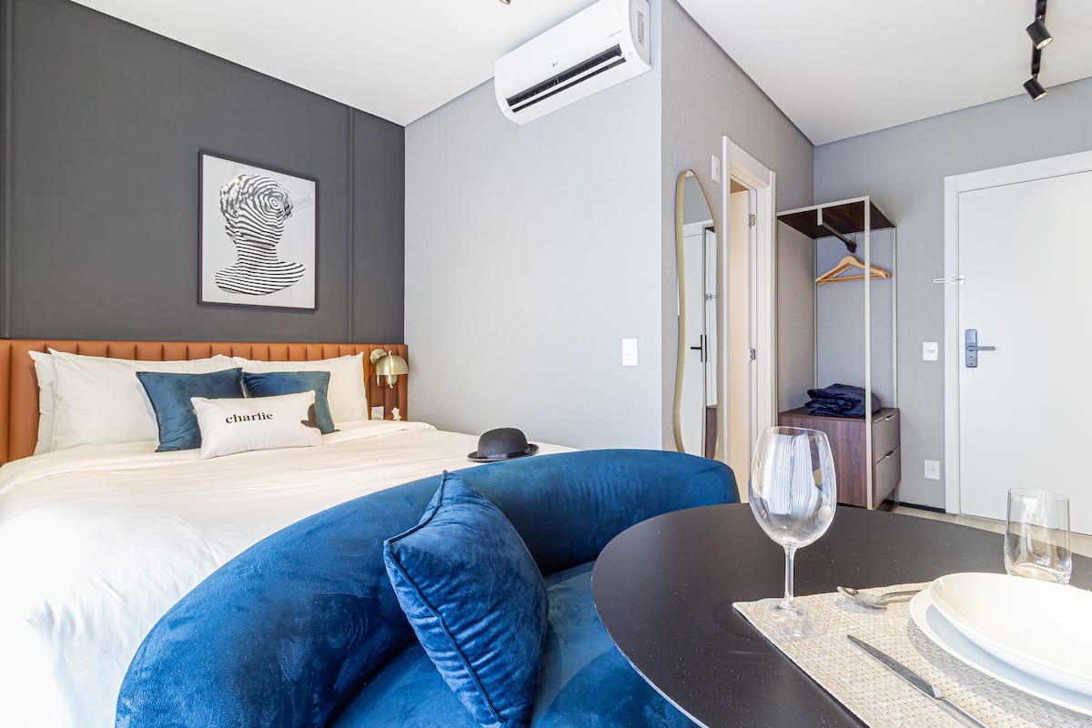 A modern hotel room with a bed, a blue sofa, a dining table with a wine glass, and a mirror.