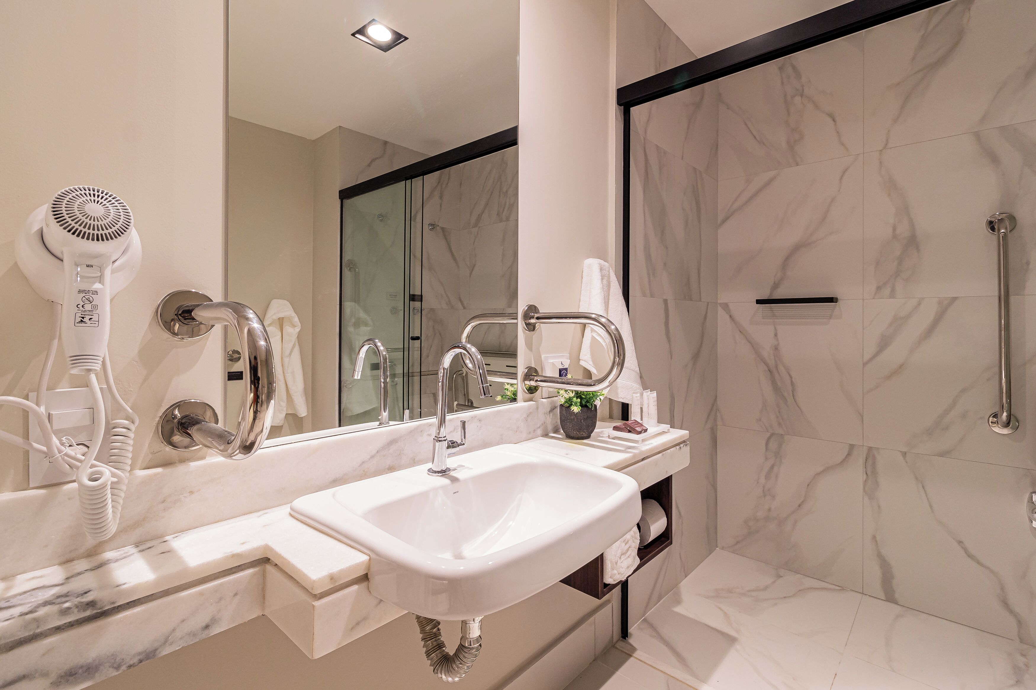 A modern bathroom with a large mirror, a white sink, a hairdryer, and a shower area.